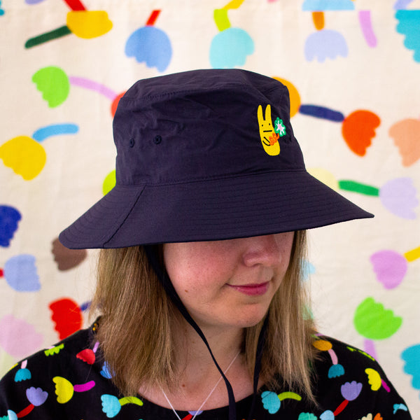 Model wearing the navy bunny bucket hat. Embroidered yellow bunny holding a green flower. 