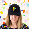 Person wearing a black baseball cap with a bright yellow bunny and green clover embroidery, standing against a colorful abstract background