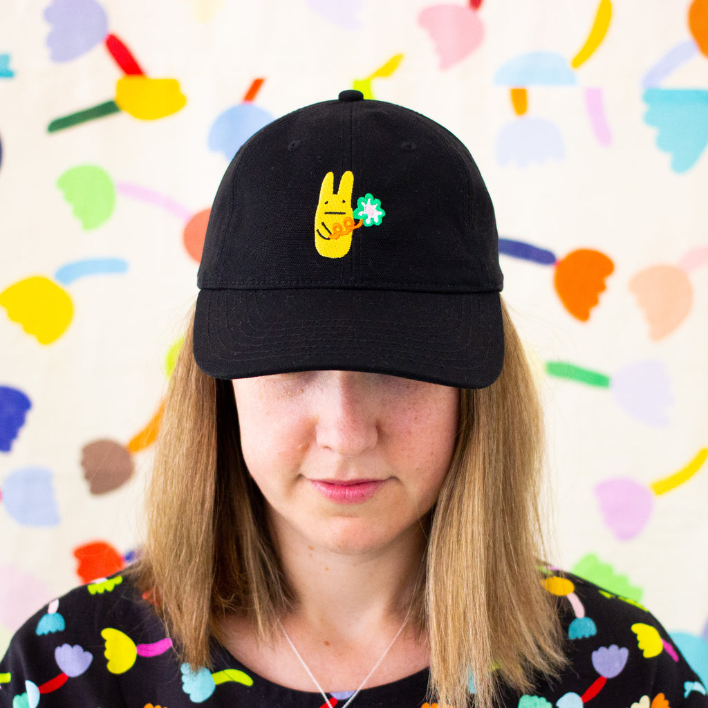 Person wearing a black baseball cap with a bright yellow bunny and green clover embroidery, standing against a colorful abstract background