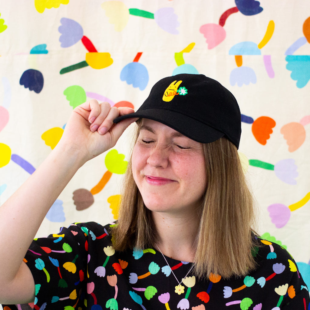 Smiling person wearing a black cap with an embroidered yellow bunny and green clover, standing in front of a colourful abstract background with eyes closed and hand on cap brim