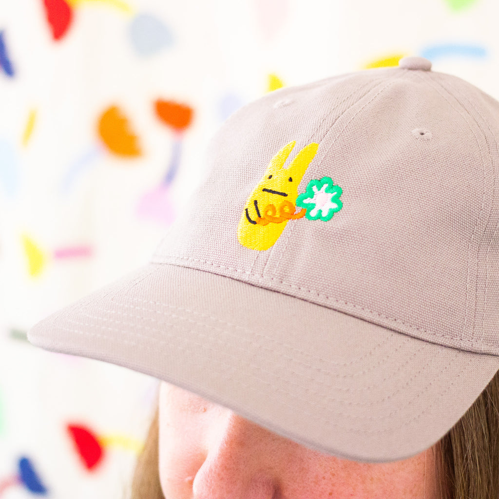 Close-up of a Mushroom coloured baseball cap featuring an embroidered yellow bunny holding a green clover, worn by a person in front of a colourful abstract background