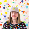 Picture of a woman wearing the bunny cap in mushroom colour. Baseball cap featuring an embroidered yellow bunny holding a green clover, worn by a person in front of a colourful abstract background