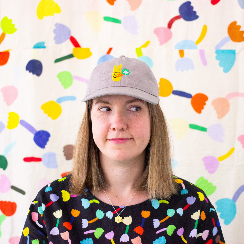 Picture of a woman wearing the bunny cap in mushroom colour. Baseball cap featuring an embroidered yellow bunny holding a green clover, worn by a person in front of a colourful abstract background