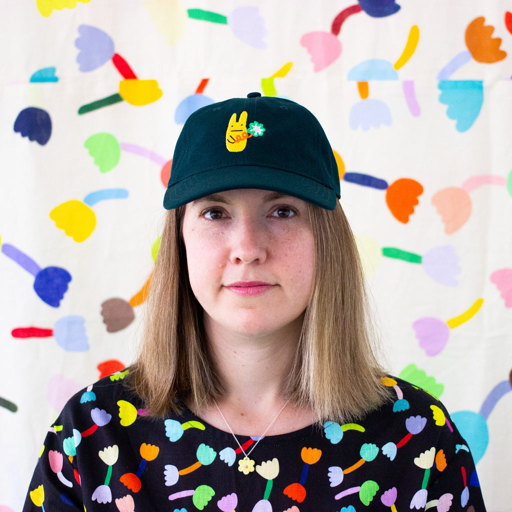 Smiling person wearing a pine green cap with an embroidered yellow bunny and green clover, standing in front of a colorful abstract background with eyes closed and hand on cap brim"