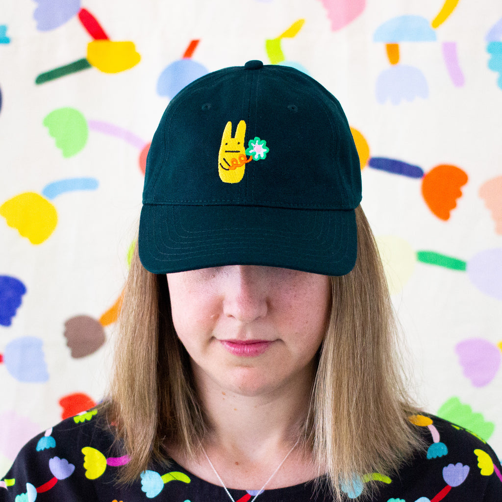 Person wearing a pine green baseball cap with a bright yellow bunny and green clover embroidery, standing against a colorful abstract background