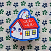 A cute cartoon house sticker with a white bunny on the red roof, placed on a patterned fabric background.