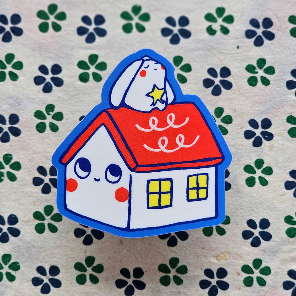 A cute cartoon house sticker with a white bunny on the red roof, placed on a patterned fabric background.