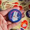 A hand holding a round blue fabric patch featuring a white bunny and the text 'tiny steps count' in pink, with a blurred background of similar Misu Juu branded patches.