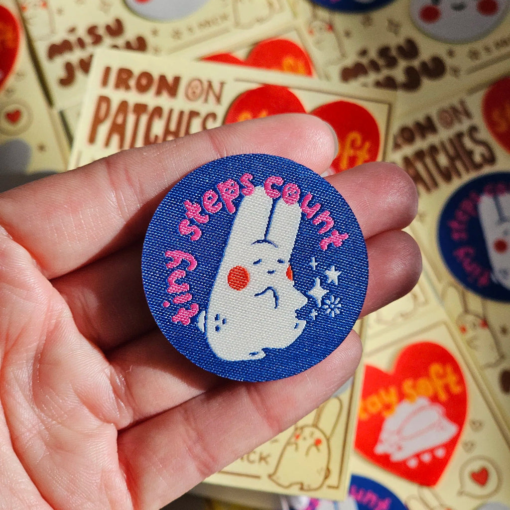 A hand holding a round blue fabric patch featuring a white bunny and the text 'tiny steps count' in pink, with a blurred background of similar Misu Juu branded patches.