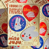 A 3-pack of Misu Juju 'Iron On Patches' featuring cute bunny and heart designs in red, blue, and white on a cream-colored backing card, surrounded by other patches.