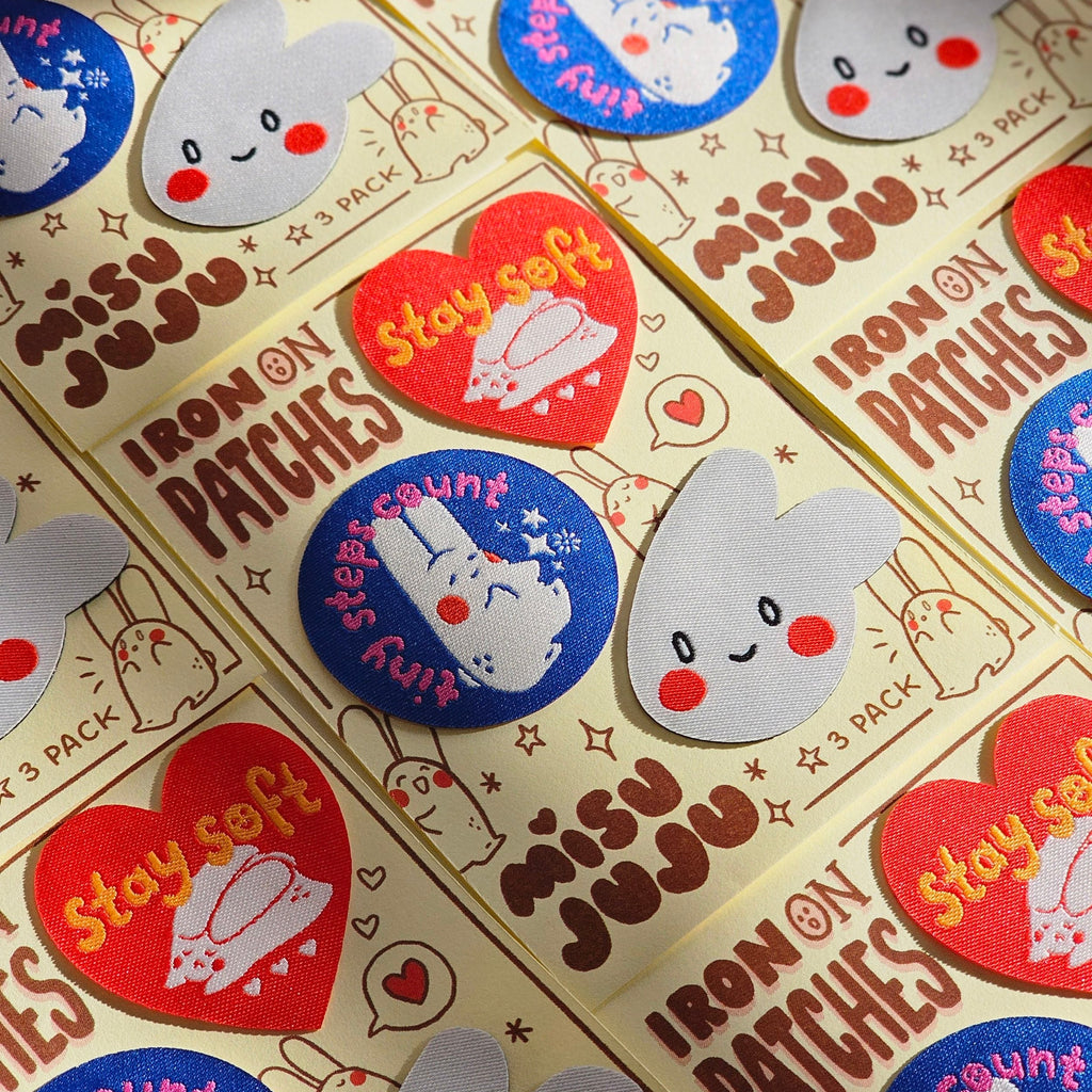 Multiple packages of Misu Juju Iron On Patches arranged diagonally. The cream-coloured packaging features brown text and cute bunny illustrations, and several patches are displayed on top, including red heart-shaped 'Stay soft' patches and blue circular 'tiny steps count' patches with white bunny designs.
