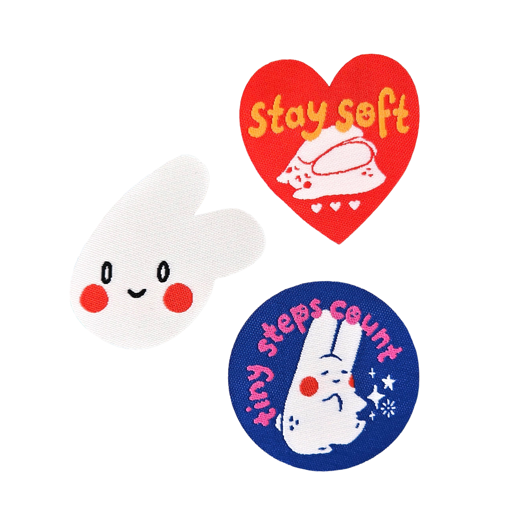 Three embroidered bunny-themed patches on a plain white background: a white bunny patch with red cheeks and a black stitched smile, a red heart patch with yellow text reading 'stay soft' and a white bunny illustration, and a blue circular patch with pink text reading 'this steps count' and a white bunny illustration.