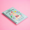 A pastel blue spiral-bound notebook with a cute bunny illustration and colourful flowers, photographed on a solid pink background.