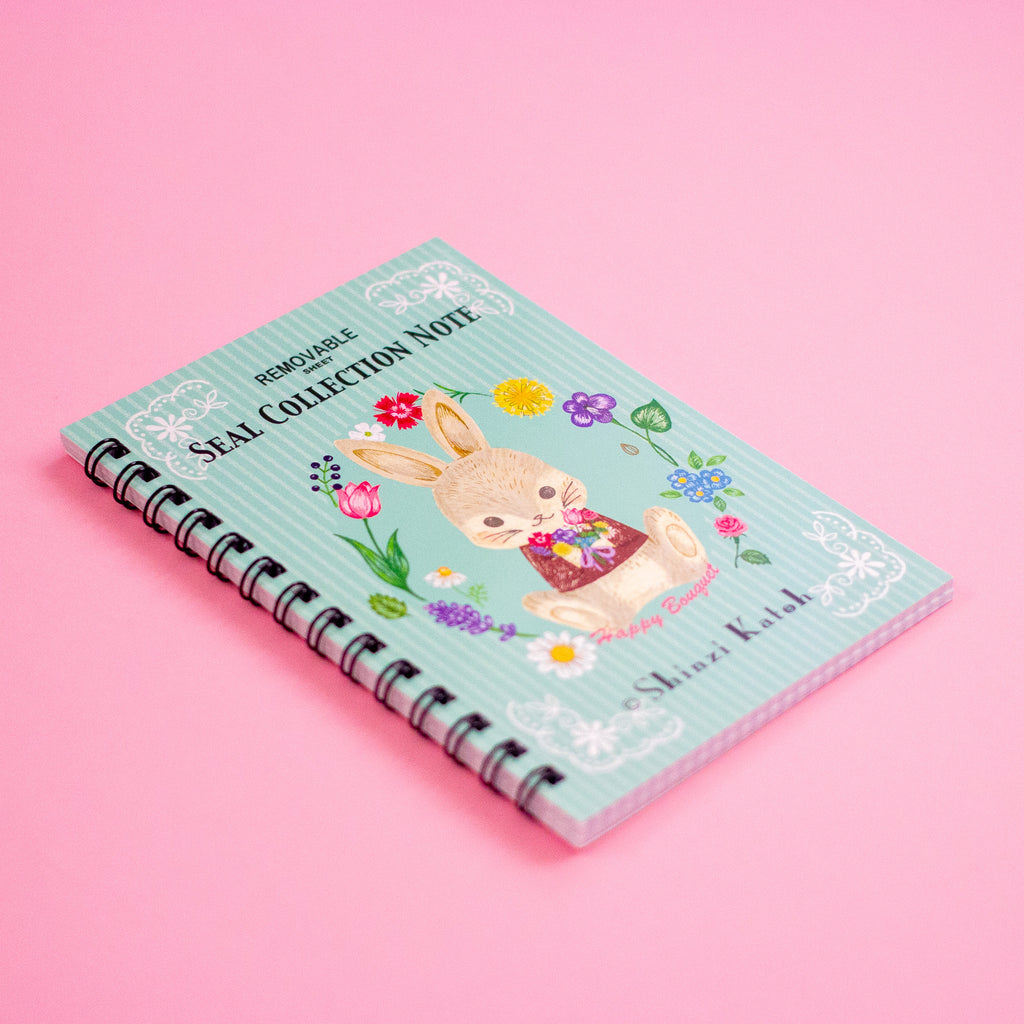 A pastel blue spiral-bound notebook with a cute bunny illustration and colourful flowers, photographed on a solid pink background.