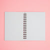 A spiral-bound notebook with a white and light blue gingham patterned cover, displayed flat against a solid pink background.