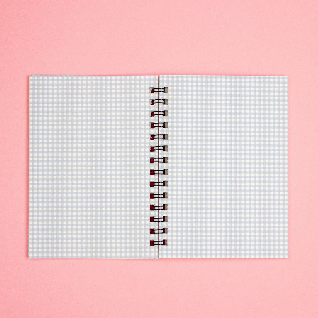 A spiral-bound notebook with a white and light blue gingham patterned cover, displayed flat against a solid pink background.