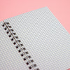A close-up of a spiral-bound notebook with a light blue and white gingham patterned cover, set against a solid pink background.