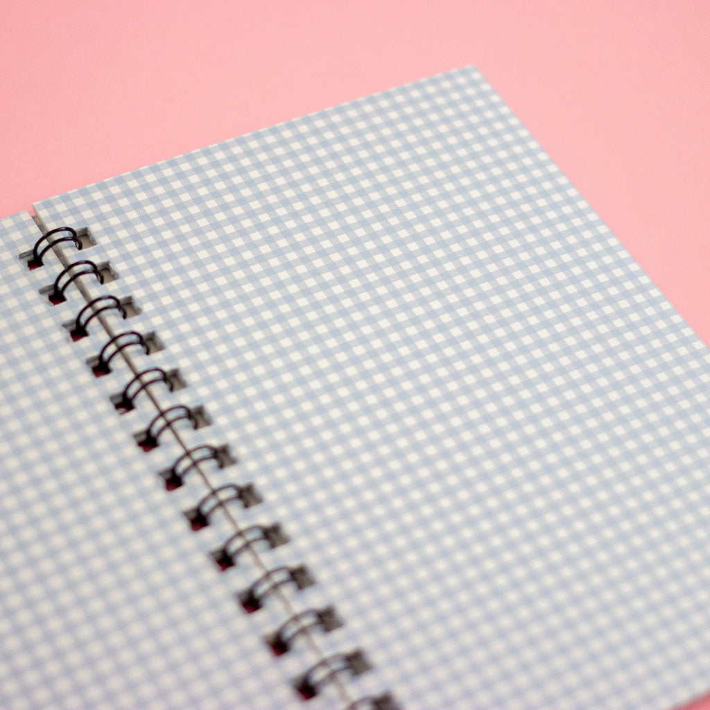 A close-up of a spiral-bound notebook with a light blue and white gingham patterned cover, set against a solid pink background.