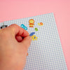 A hand peeling a small colourful sticker from a light blue and white gingham notepad on a solid pink background.