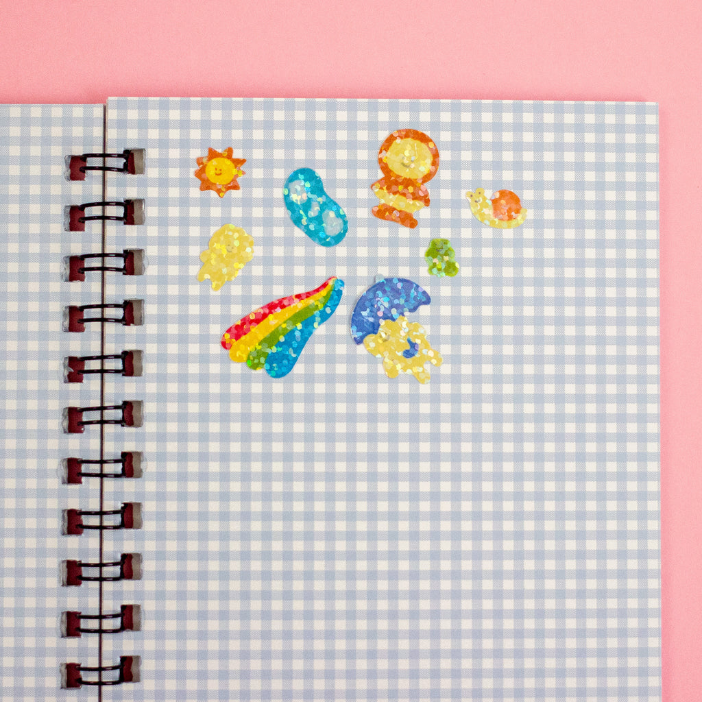 A spiral-bound notebook with a light blue and white gingham cover, decorated with colourful glittery stickers against a solid pink background.