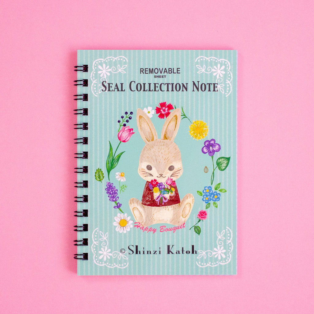 A spiral-bound notebook titled 'Removable Seal Collection Note' by Shinzi Katoh, featuring a cute bunny illustration on a pastel blue striped cover, set against a solid pink background.