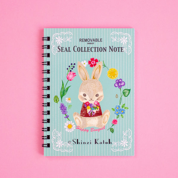 A spiral-bound notebook titled 'Removable Seal Collection Note' by Shinzi Katoh, featuring a cute bunny illustration on a pastel blue striped cover, set against a solid pink background.