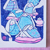 A square art print featuring a whimsical illustration of rabbits wrapped in patterned blankets, with a teapot and pink bedding, shown against a dark blue background with a white border. The print is photographed standing on a pink fabric surface.