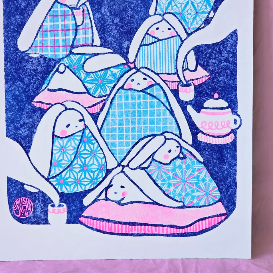 A square art print featuring a whimsical illustration of rabbits wrapped in patterned blankets, with a teapot and pink bedding, shown against a dark blue background with a white border. The print is photographed standing on a pink fabric surface.