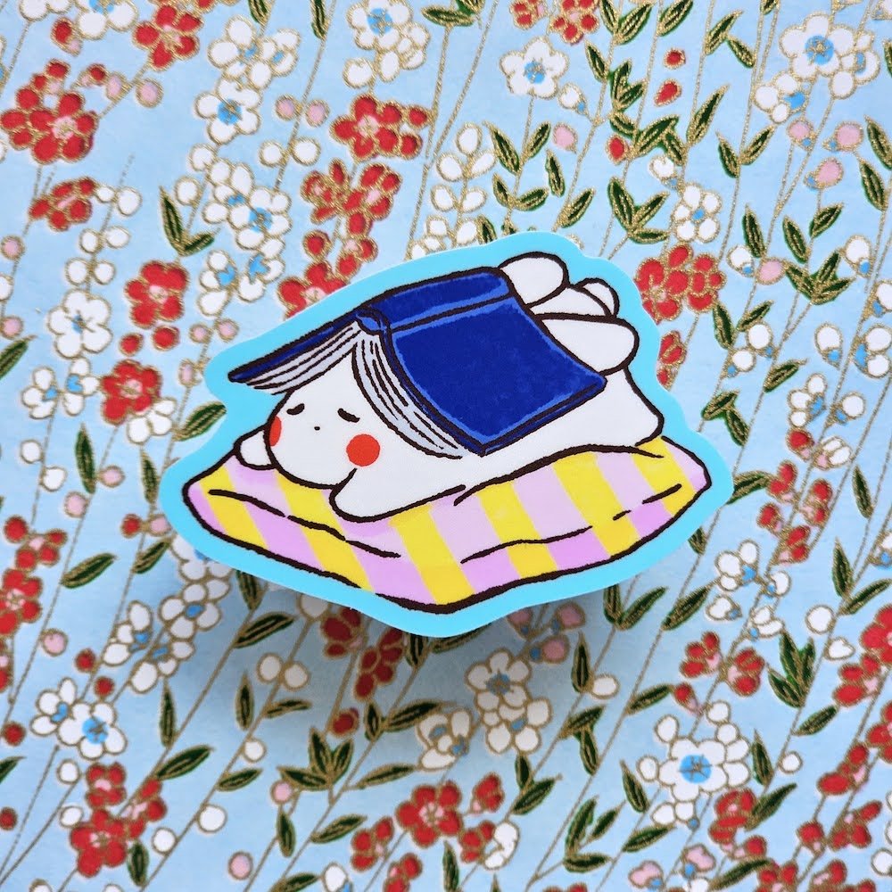 A cute cartoon sticker of a white bunny wearing a blue cap and lying on a pink and yellow striped blanket, outlined in bright blue, shown on a floral background.