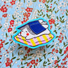 A cute cartoon sticker of a white bunny wearing a blue cap and lying on a pink and yellow striped blanket, outlined in bright blue, shown on a floral background.