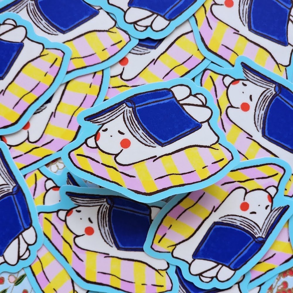 A pile of colourful die-cut stickers featuring a cartoon illustration of a white bunny under an open book, on a pink and yellow striped cushion. The stickers have a bright blue border.