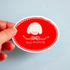 A hand holding a red oval sticker with a white scalloped border, a small white sleeping character, and the text 'busy dreaming' against a light blue background.