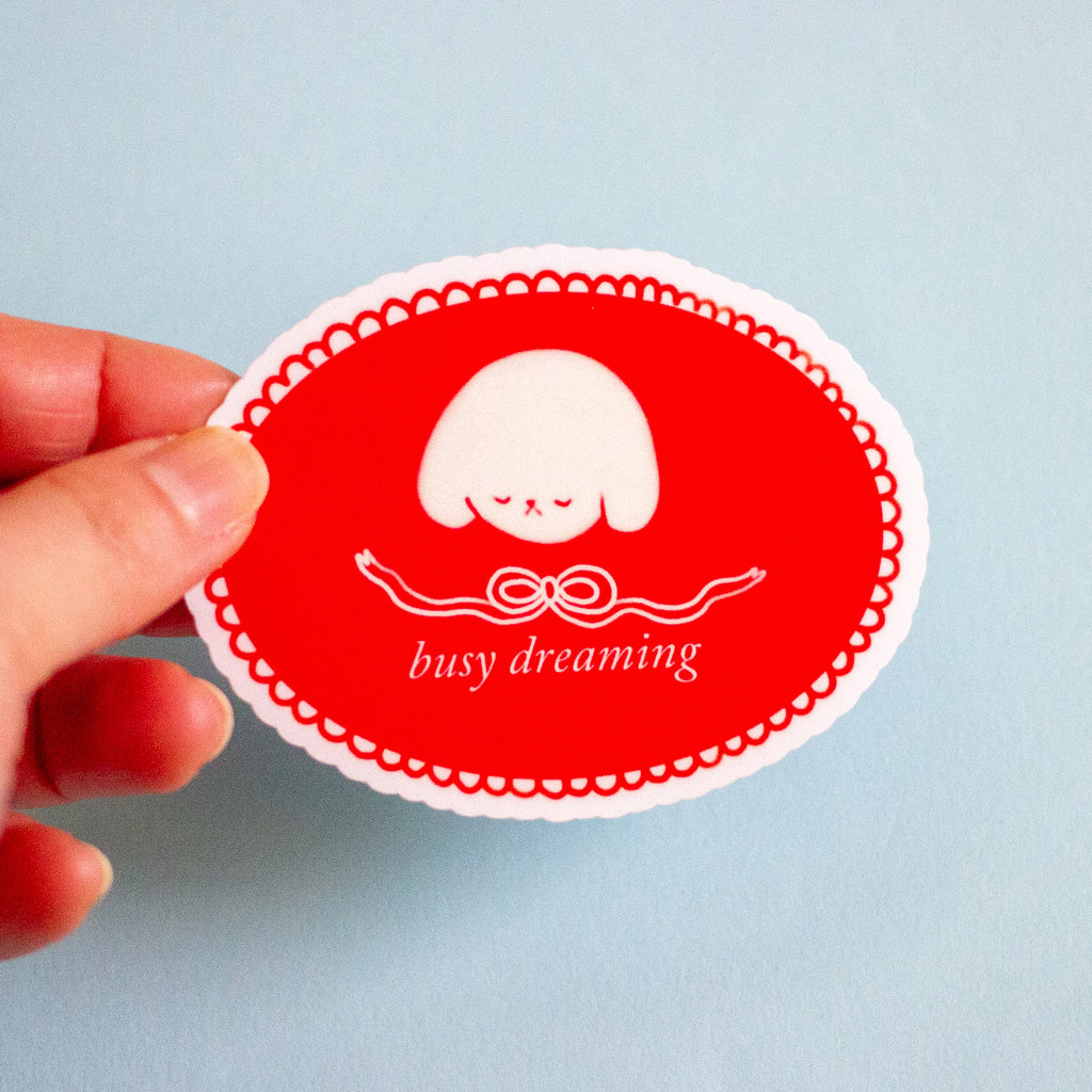A hand holding a red oval sticker with a white scalloped border, a small white sleeping character, and the text 'busy dreaming' against a light blue background.