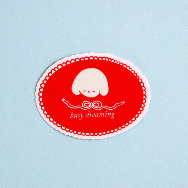 An oval red sticker with a scalloped white border, featuring a white sleeping cartoon character and the text 'busy dreaming' on a light blue background.
