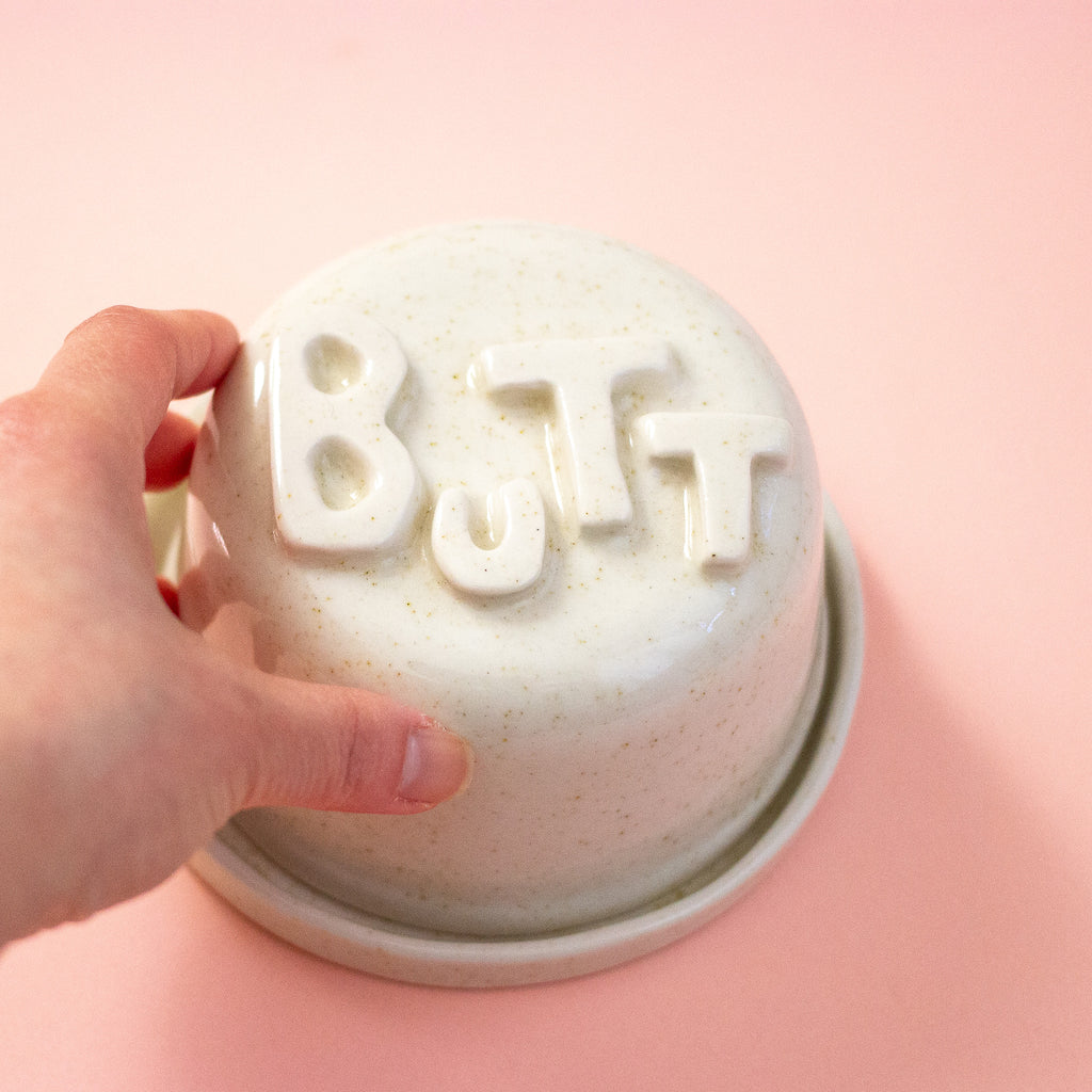 Hand holding a ceramic container with 'BUTT' on a peach background
