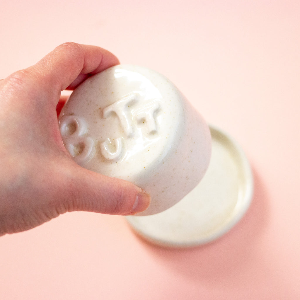 Hand holding a lid with 'BUTT' letters against a peach background