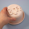 Hand pressing a pink lid with 'BUTT' letters on a light blue background