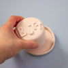 Hand holding a pink object with 'BUTT' printed on it against a blue background