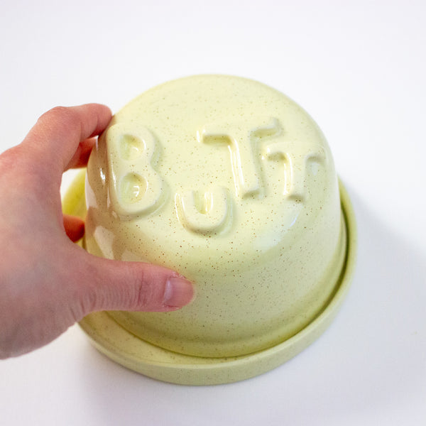 Hand holding a light yellow ceramic bowl with 'BUTT' embossed on it against a white background
