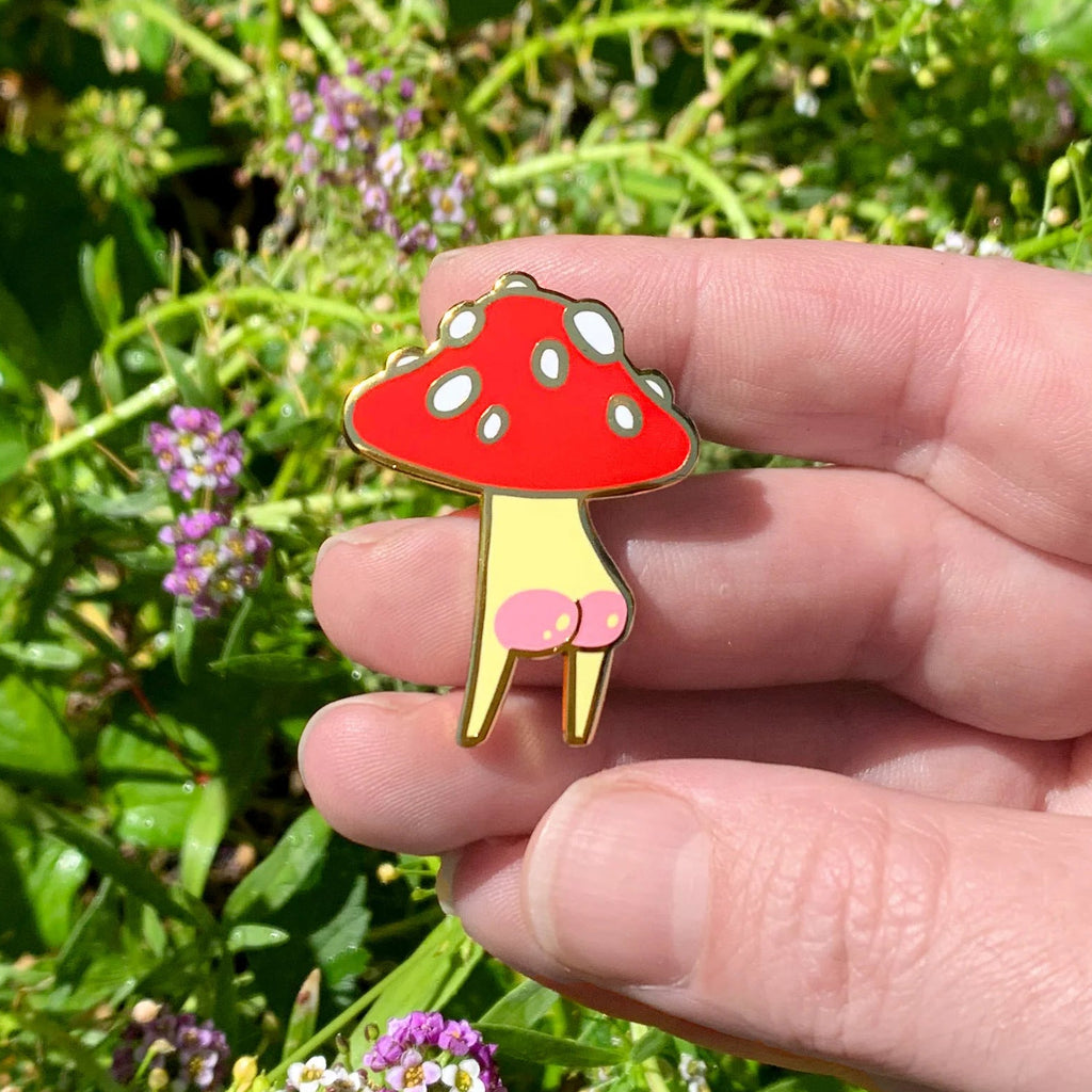 Hand holding a red and gold mushroom-shaped pin against a natural background with greenery and flowers.