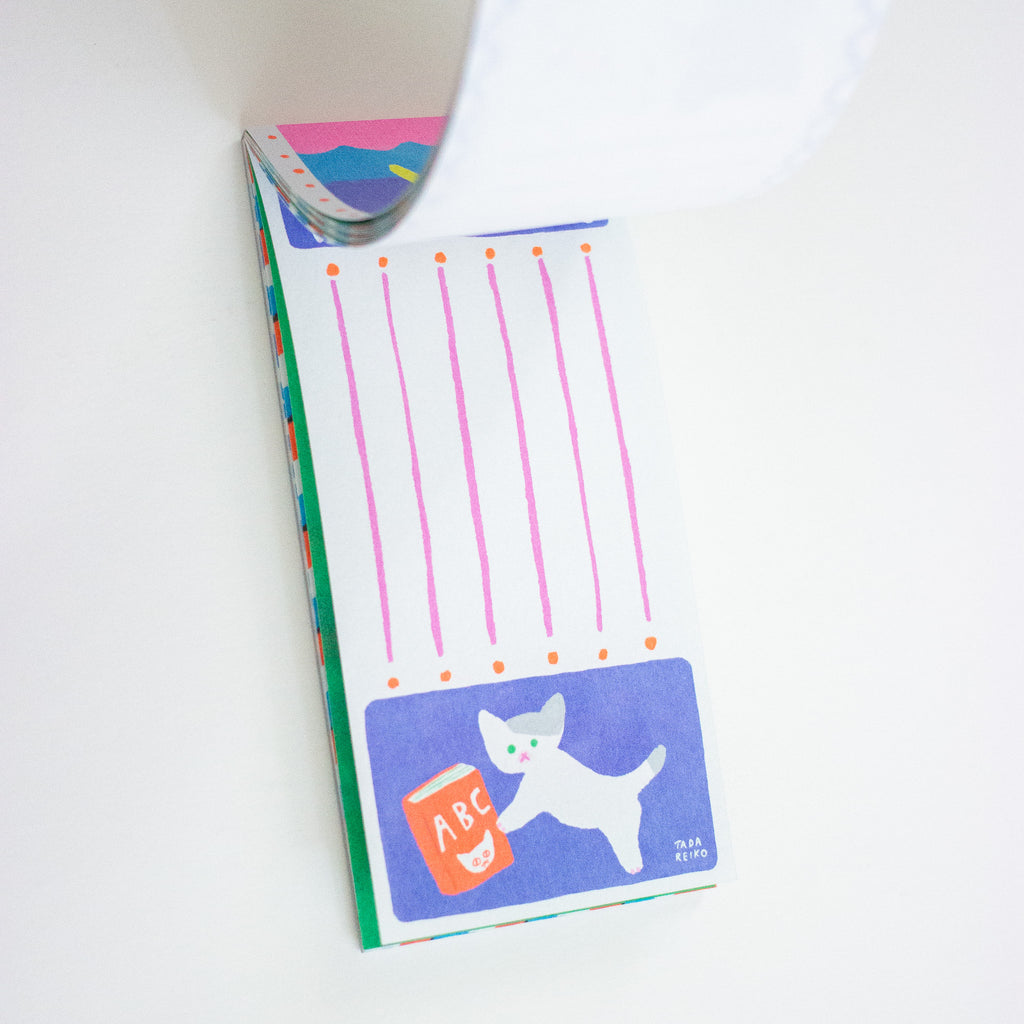 A close-up of a colourful printed notepad on a white background, showing pink vertical lines, small orange dots, and a blue panel with a white cat holding an orange book labeled 'ABC'.