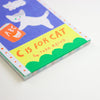 A close-up of the book cover of 'C Is For Cat' by Tada Reiko, featuring a playful, colourful illustration of a white cat on a blue background with a yellow banner and green scallop border.