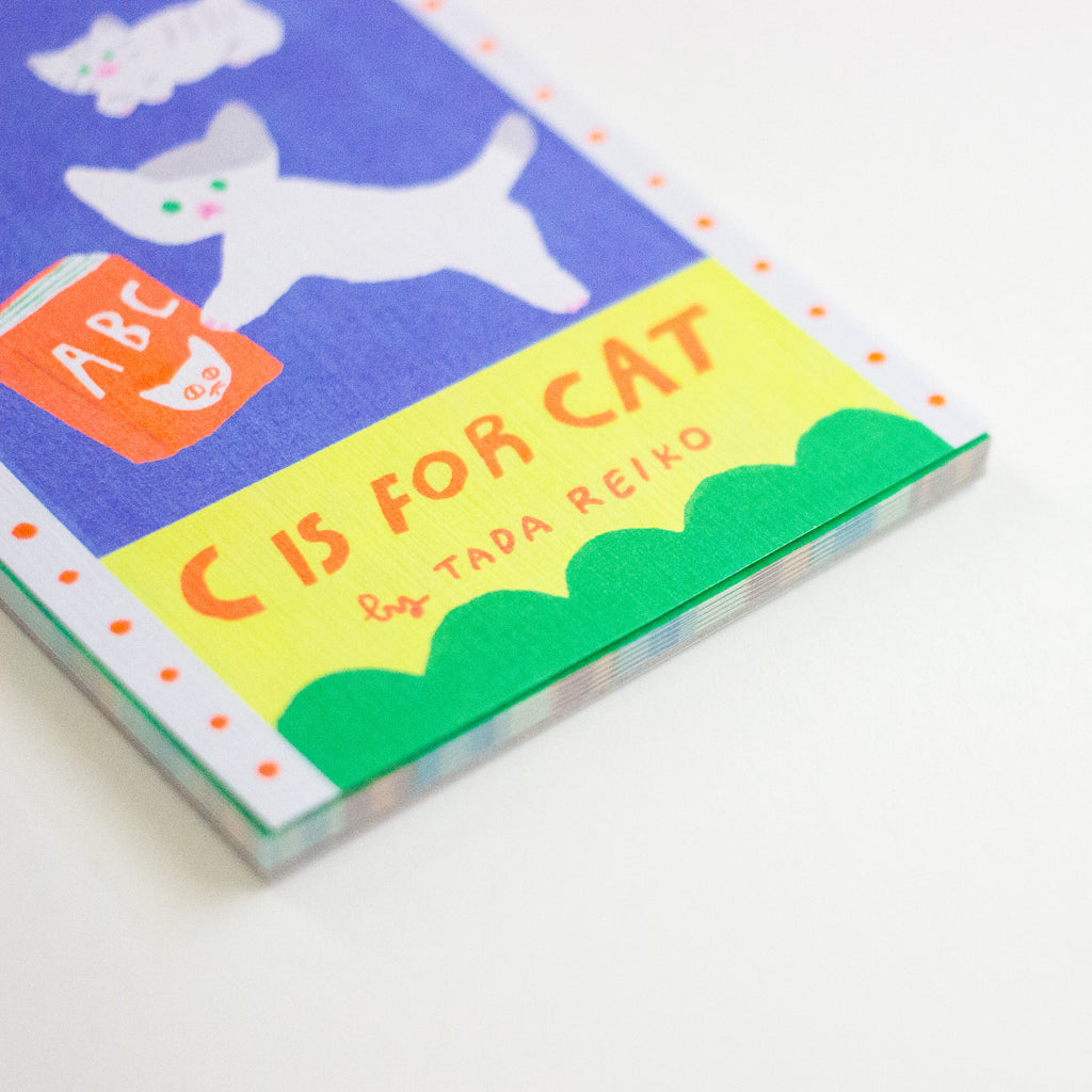A close-up of the book cover of 'C Is For Cat' by Tada Reiko, featuring a playful, colourful illustration of a white cat on a blue background with a yellow banner and green scallop border.