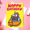 A yellow birthday card with a cartoon cake slice and a cat, reading 'HAPPY BIRTHDAY!' in pink letters, styled on a pink background with white envelopes, markers, binder clips, and flowers.