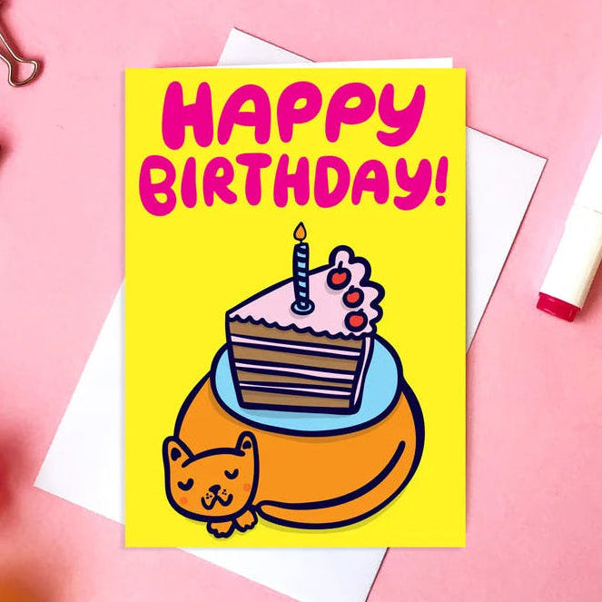 A yellow birthday card with a cartoon cake slice and a cat, reading 'HAPPY BIRTHDAY!' in pink letters, styled on a pink background with white envelopes, markers, binder clips, and flowers.