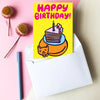 A yellow birthday card with a cartoon cake slice and a cat, reading 'HAPPY BIRTHDAY!' in pink letters, styled on a pink background with white envelopes, markers, binder clips, and flowers.