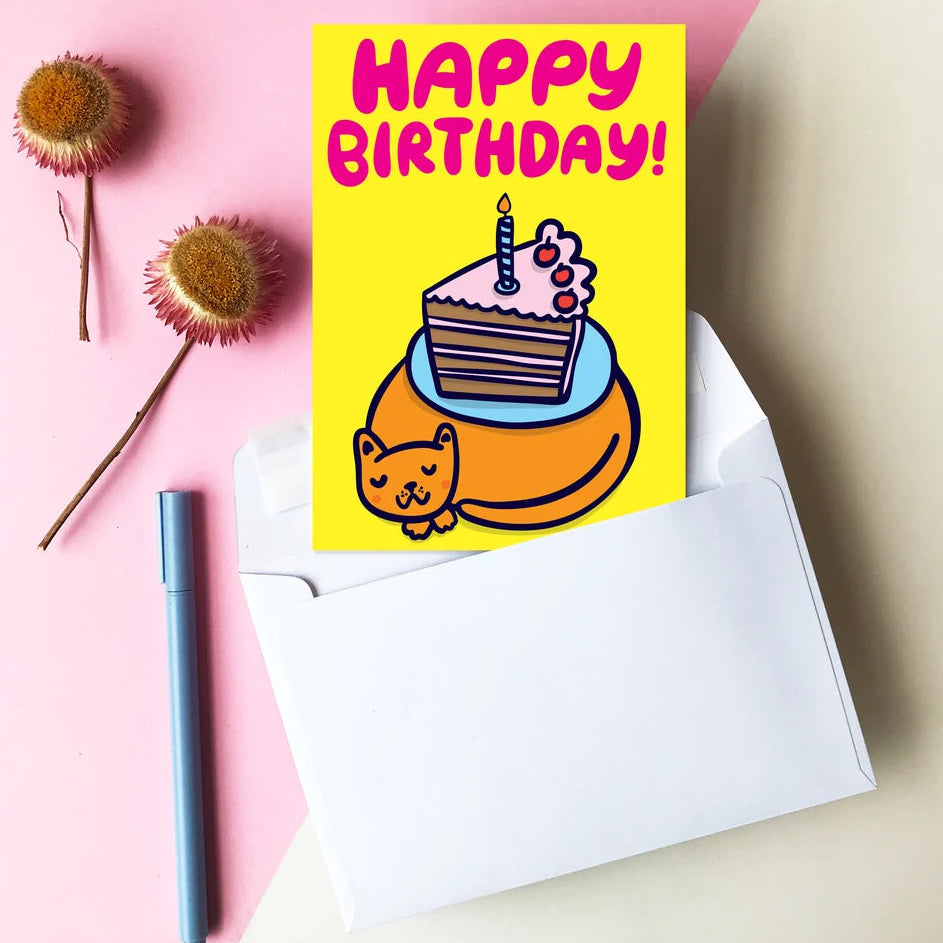 A yellow birthday card with a cartoon cake slice and a cat, reading 'HAPPY BIRTHDAY!' in pink letters, styled on a pink background with white envelopes, markers, binder clips, and flowers.