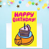 A yellow birthday card with bold pink text reading 'HAPPY BIRTHDAY!' and a cartoon cake slice with a candle and a sleeping cat, shown standing on a white envelope against a light blue background with confetti and star decorations.