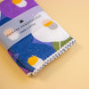 A folded Japanese handkerchief with a colourful abstract pattern in purple, blue, white, yellow, and green, wrapped in a clear paper band labeled 'HAIKARA HANDKERCHIEF made in JAPAN', on a solid pale yellow background.