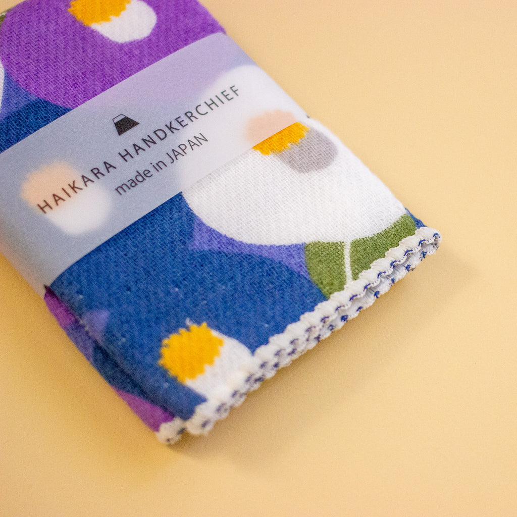 A folded Japanese handkerchief with a colourful abstract pattern in purple, blue, white, yellow, and green, wrapped in a clear paper band labeled 'HAIKARA HANDKERCHIEF made in JAPAN', on a solid pale yellow background.